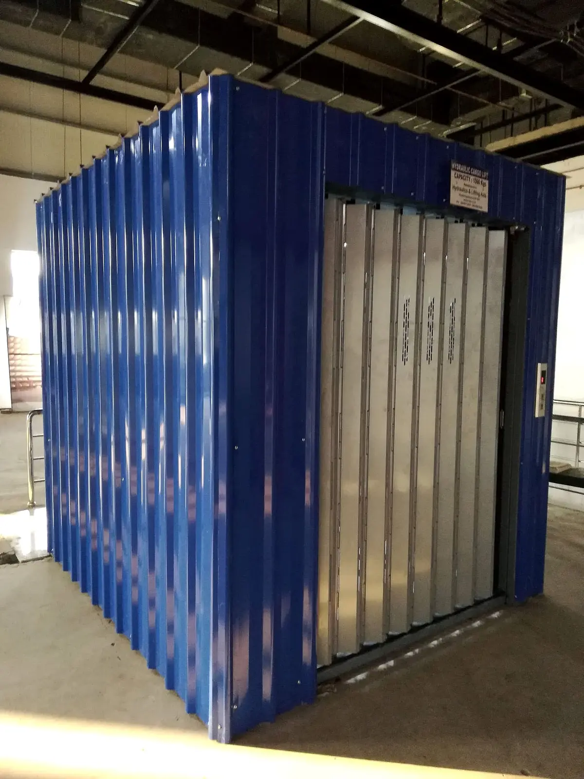 Hydraulic goods lift in a blue steel enclosure with a folding gate
